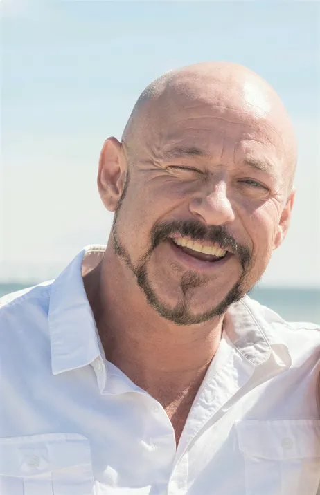 A bald man with a goatee smiles and winks at the camera, wearing a white shirt with a beach and blue sky in the background.