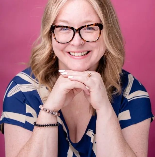A person with blonde hair and glasses smiles, resting their hands under their chin, wearing a blue patterned top against a pink background.