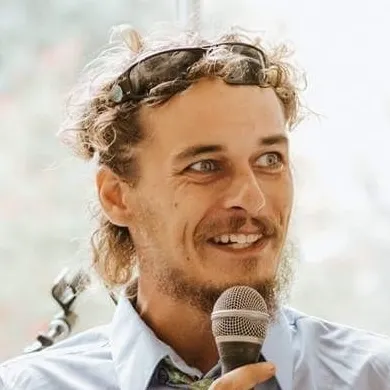 A man with curly hair and a beard, wearing sunglasses on his head and a light blue shirt, holds a microphone and speaks, smiling slightly.